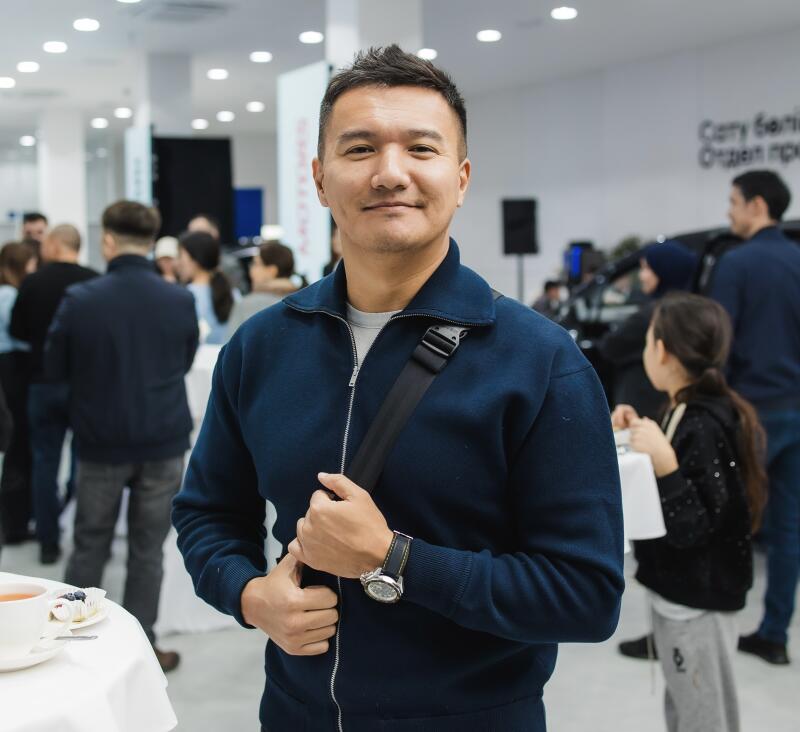 A man in a blue jacket with a strap over his shoulder stands in a room with other people and furniture.