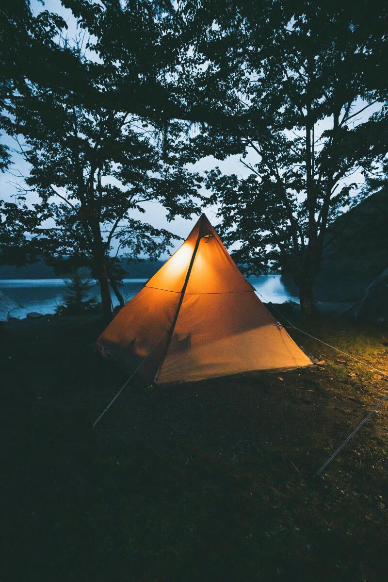 A glowing orange tent is set up on a grassy area surrounded by trees, with a body of water visible in the background.