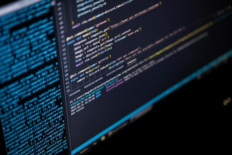 A monitor displays code in a dark theme, with a mix of Python and other script languages visible. The code includes URLS, function definitions, and module imports, alongside system information and timestamps at the bottom.