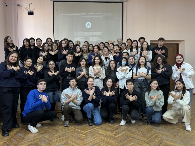 A group of about 40 young people pose for a photo in a classroom, all making the same hand gesture by crossing their hands over their chests; a projector screen is visible in the background.
