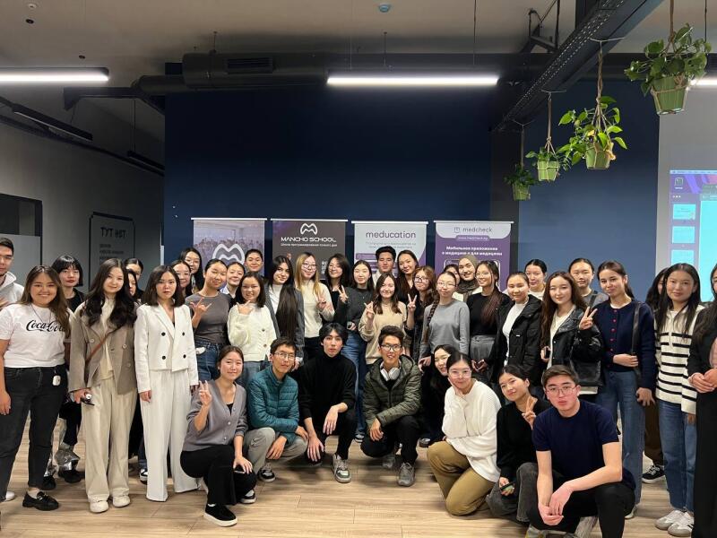 A group of young people are posing for a group photo in a room with banners for 'MANCHO SCHOOL,' 'meducation,' and 'medcheck' in the background.