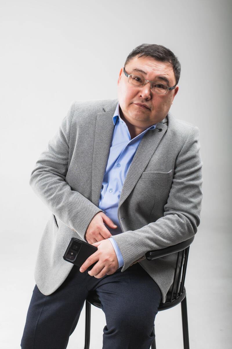 A middle-aged man with glasses, wearing a grey blazer and a light blue shirt, is sitting on a chair and holding a smartphone.
