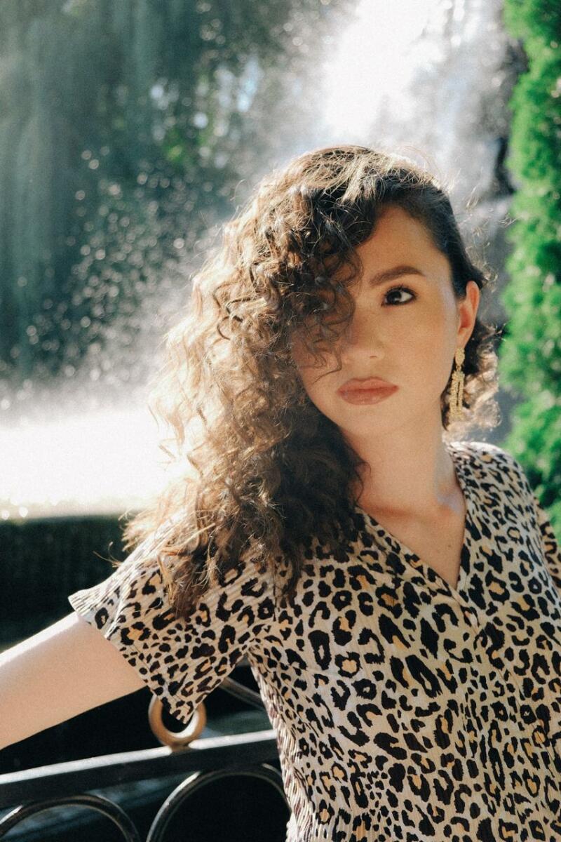 A woman with curly hair is wearing a leopard-print shirt and dangling earrings, posing in front of a blurred fountain.