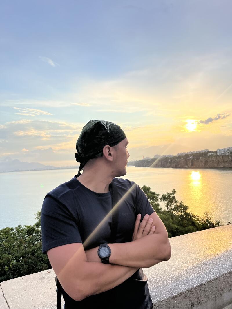 A man with a headscarf and a watch stands with his arms crossed, looking out over the water towards the sunset.