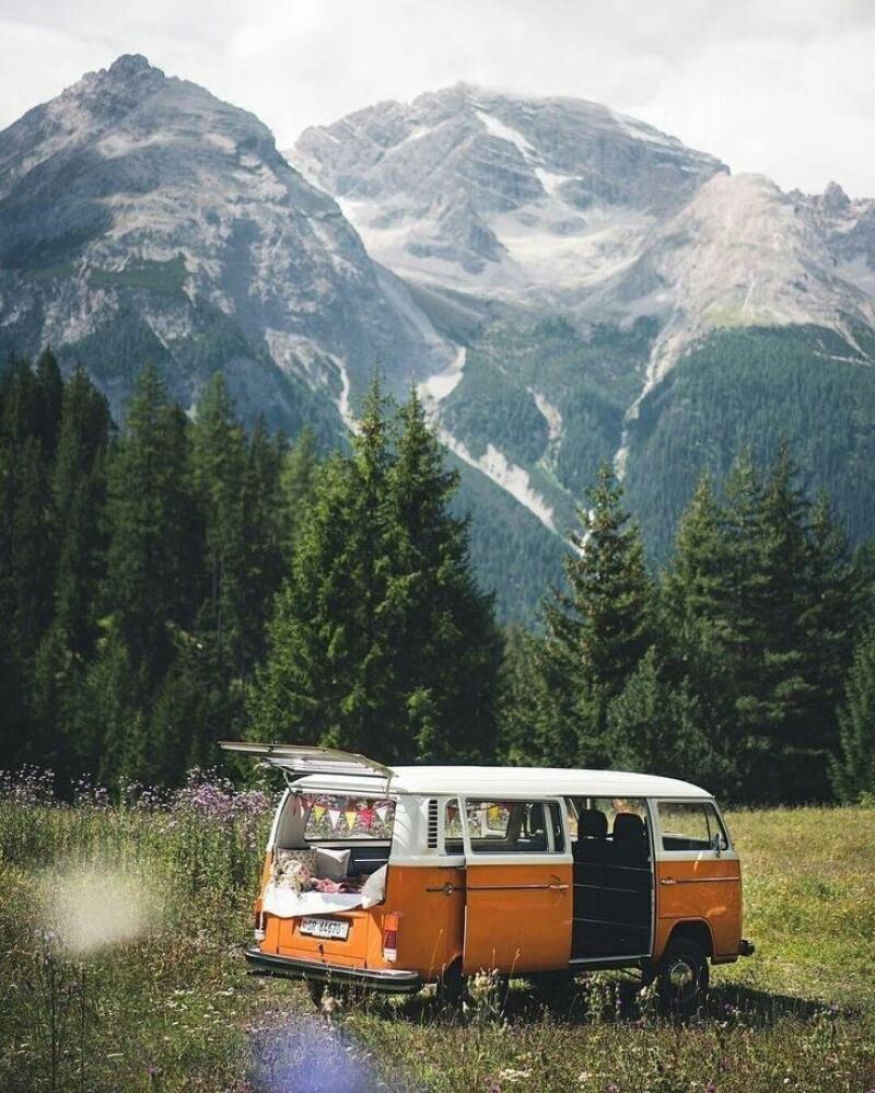 An orange and white Volkswagen van is parked in a field with the back door open, revealing a bed inside, with tall mountains and trees in the background.