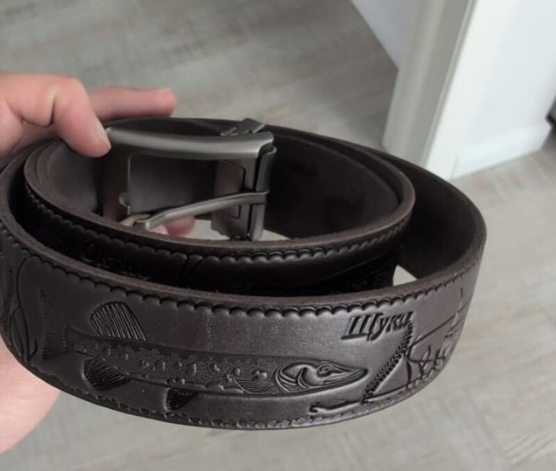 A brown leather belt with an embossed design of a pike fish and the word "щука" (pike) in Russian, featuring a silver-colored buckle, held in a hand.