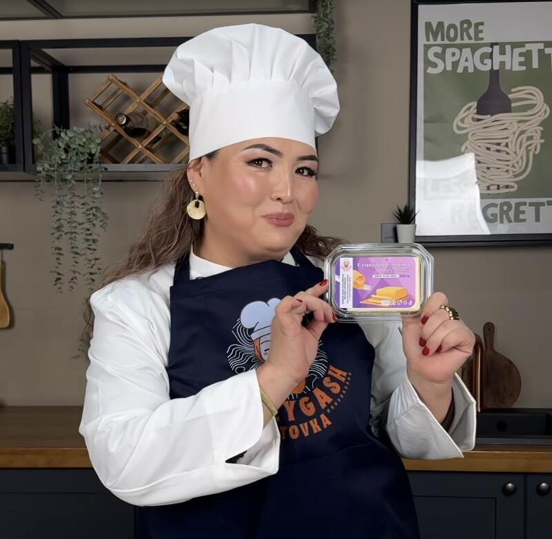 A woman in a chef's hat and apron is holding a package of cheese, with a poster that says "More Spaghetti Less Regretti" in the background.