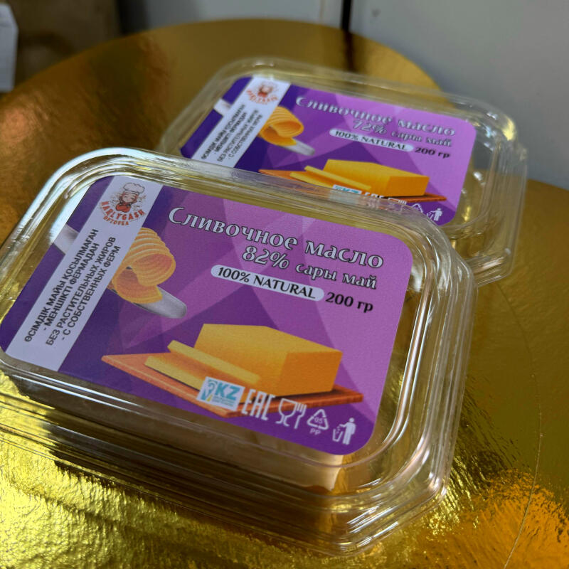 Two clear plastic containers with labels for "Slivochnoye Maslo" (butter) are stacked on a gold surface; the top container reads "72%" and the bottom one reads "82%" with the text "100% NATURAL 200 гр" on both.