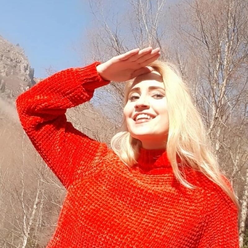 A smiling woman with blonde hair wearing a red sweater shields her eyes with her hand against a backdrop of trees and mountains.