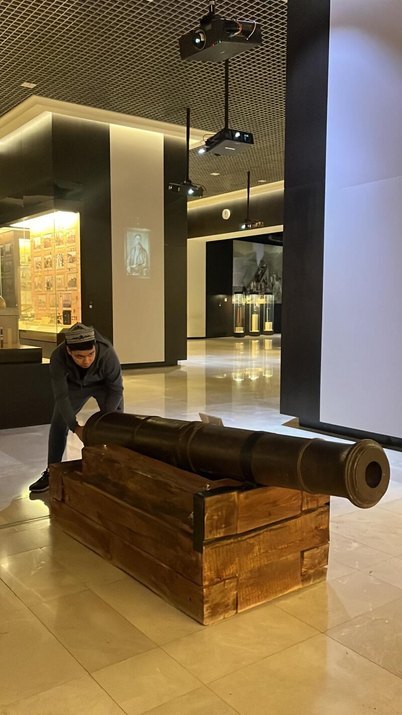 A man in a skullcap and gray suit pushes a cannon mounted on a wooden stand in a museum with projectors on the ceiling.