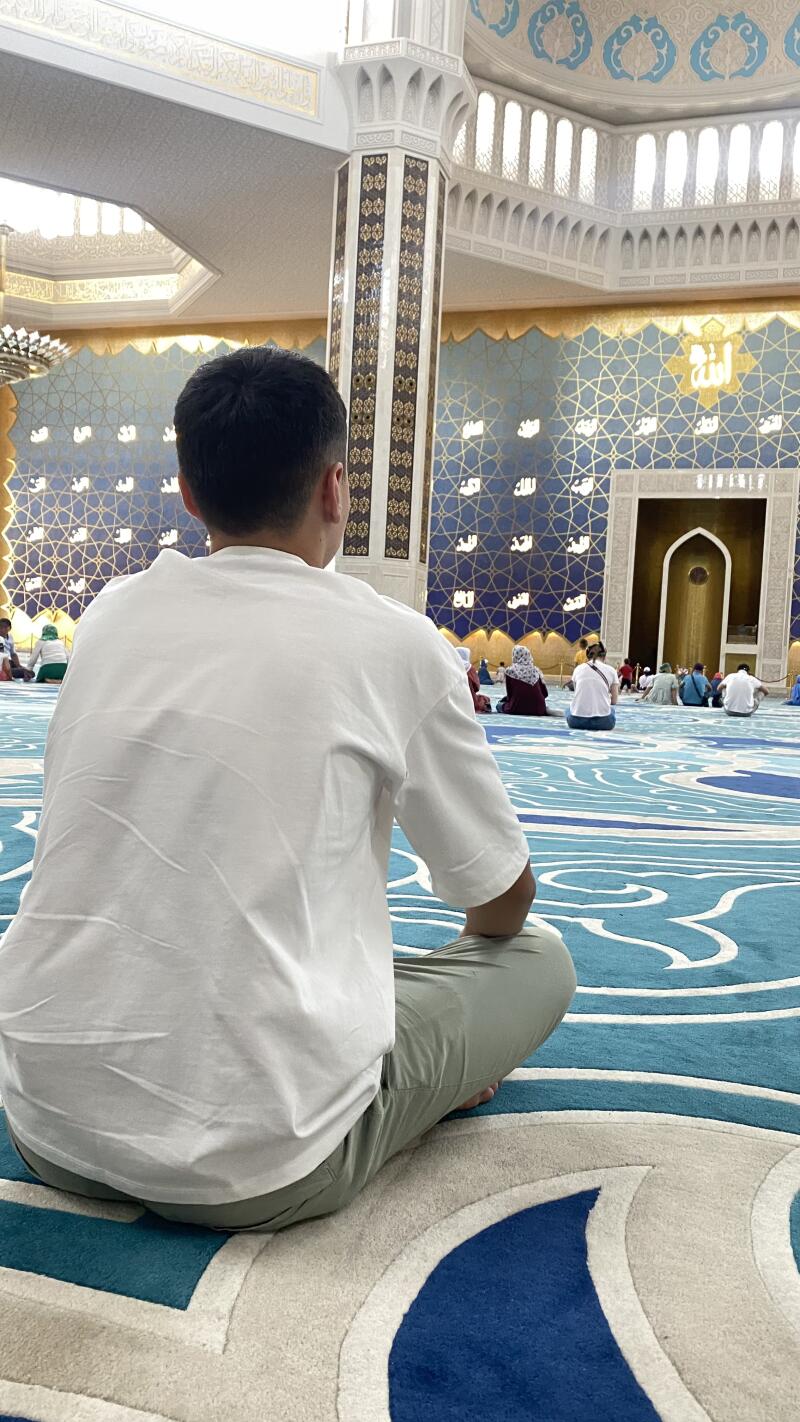 A man wearing a white t-shirt and green pants is sitting in a mosque with blue carpet and ornate walls, with the word "Allah" visible on the wall.
