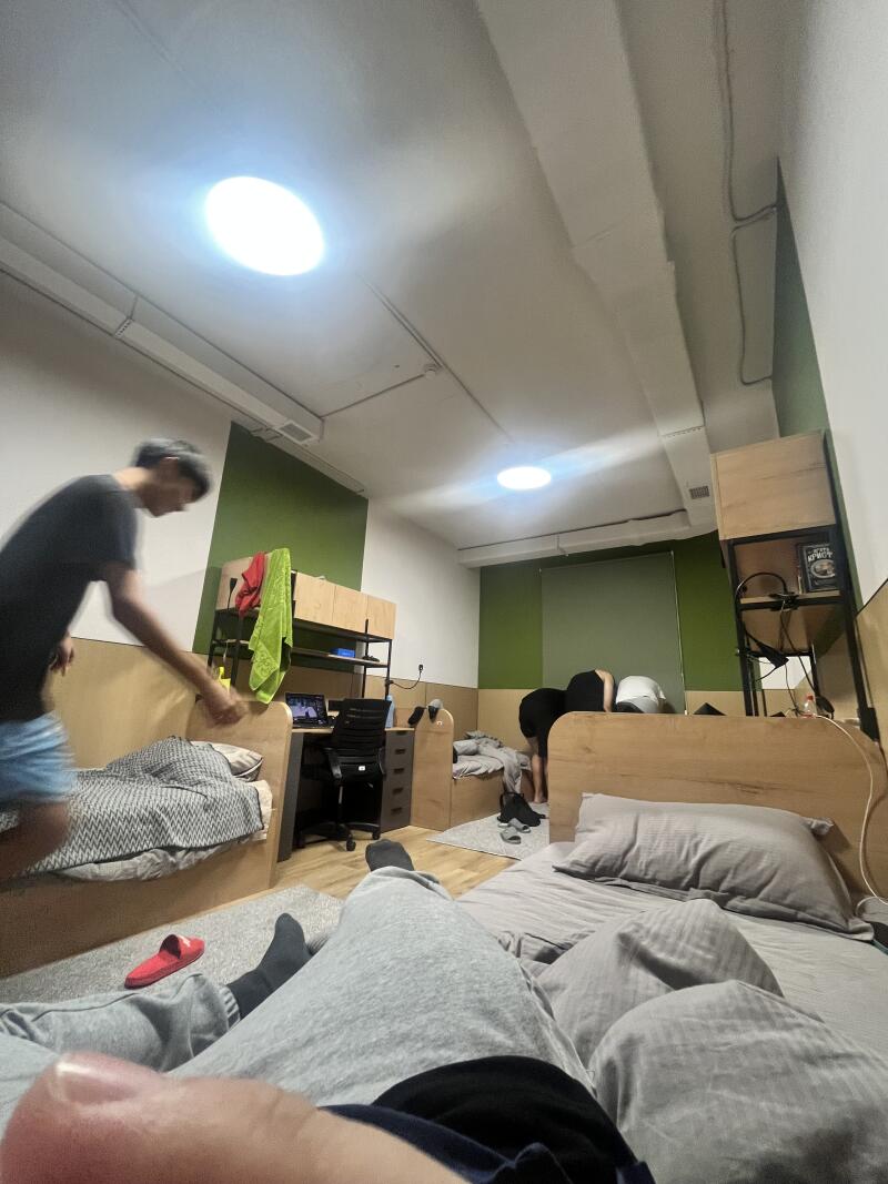 A dorm room is shown with two people standing near separate beds, while a person is lying in bed in the foreground.