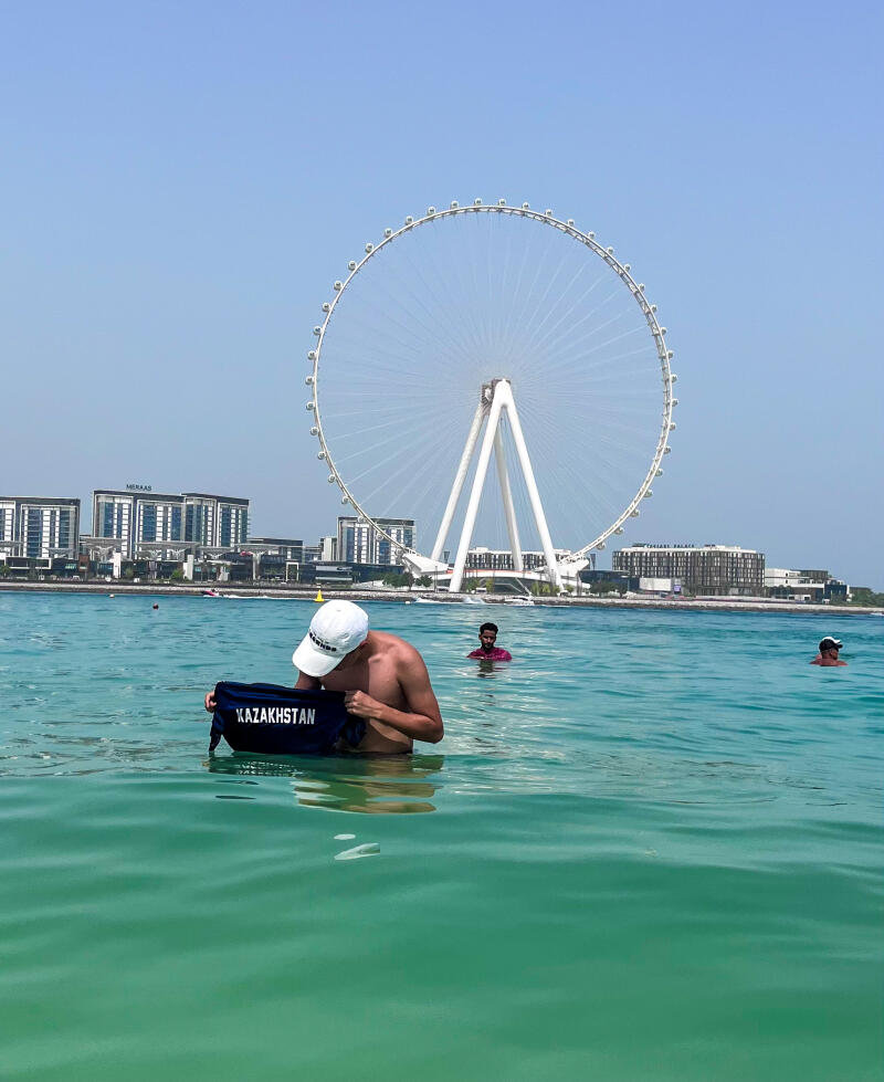 A shirtless man in a white cap stands waist-deep in turquoise water, holding a dark blue bag with the word "KAZAKHSTAN" printed on it; in the background, there is a large Ferris wheel and buildings under a clear sky.
