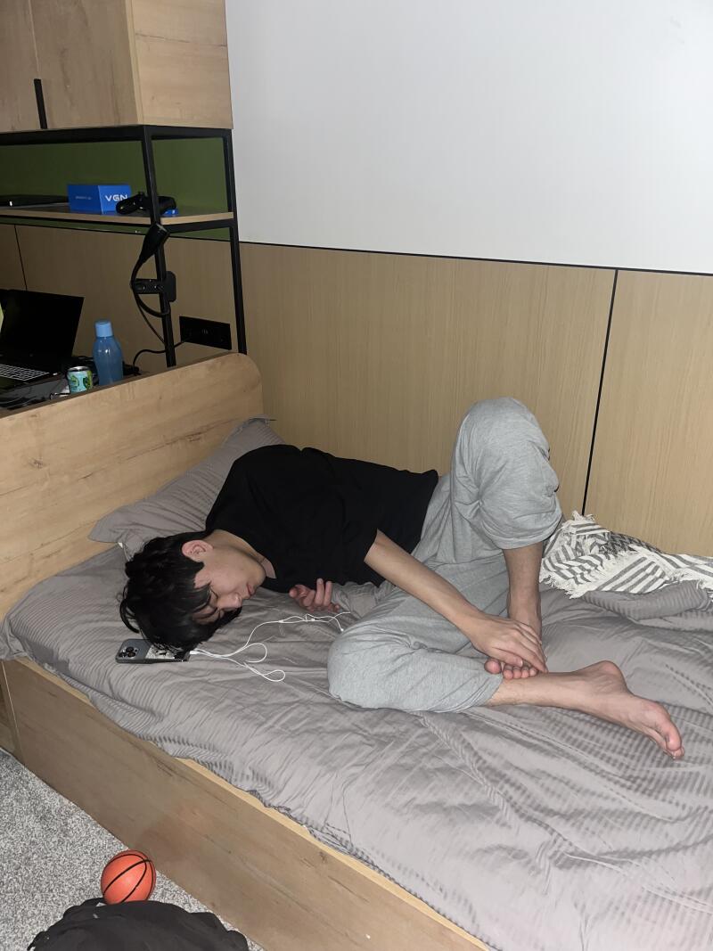 A person in a black shirt and gray sweatpants lies on a bed with a gray comforter, with a phone and white earphones next to them.