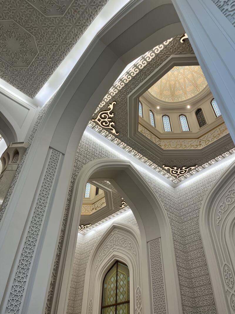The image shows the interior of a mosque, featuring white arches and walls adorned with intricate patterns and designs, leading up to a dome with windows.
