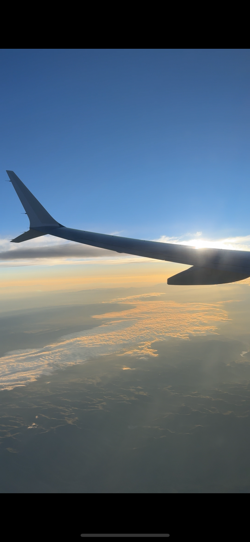 The photo shows a view from an airplane window, with the wing of the plane visible against a backdrop of blue sky and clouds, and the sun is shining.