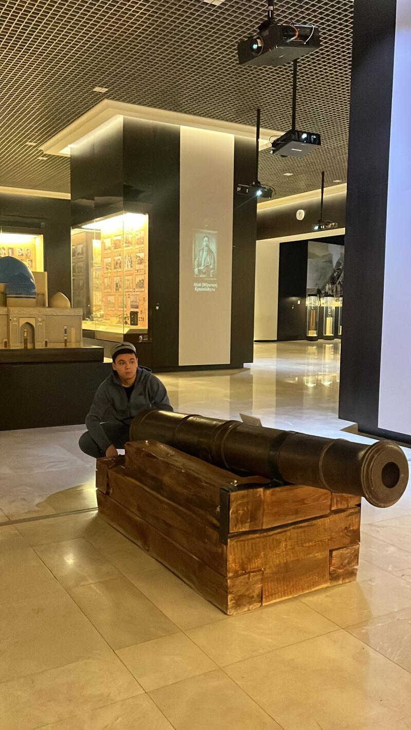 A person in a hat crouches next to a cannon on a wooden base, inside a museum with displays and projectors on the ceiling.