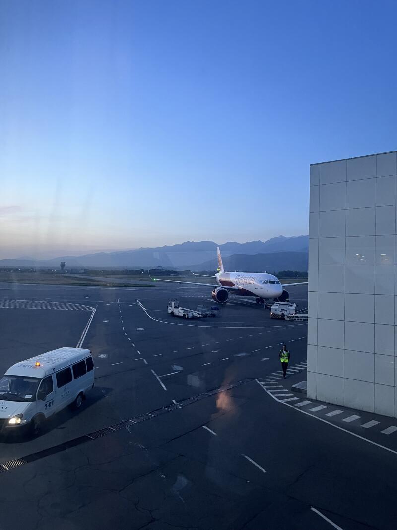 The photo shows an airplane parked on an airport tarmac with a white van and a person in a reflective vest nearby, under a blue sky with mountains in the background.