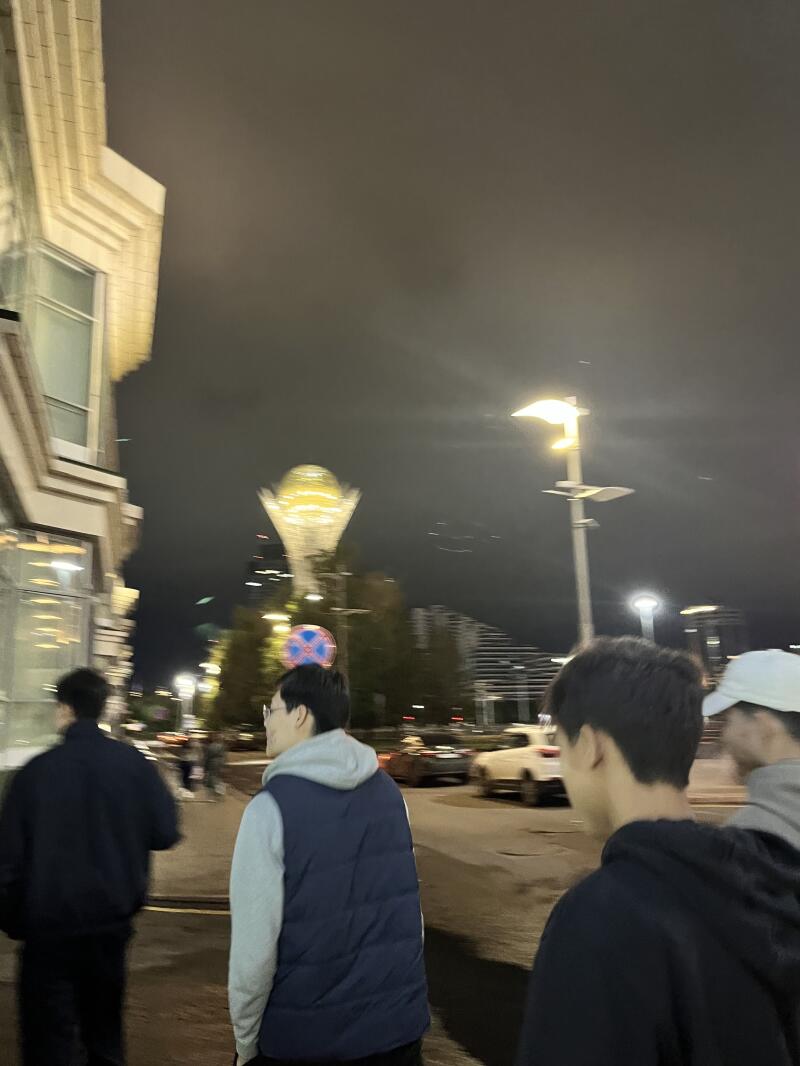 At night, a group of young men are walking down a city street with illuminated buildings and the Bayterek monument in the background.