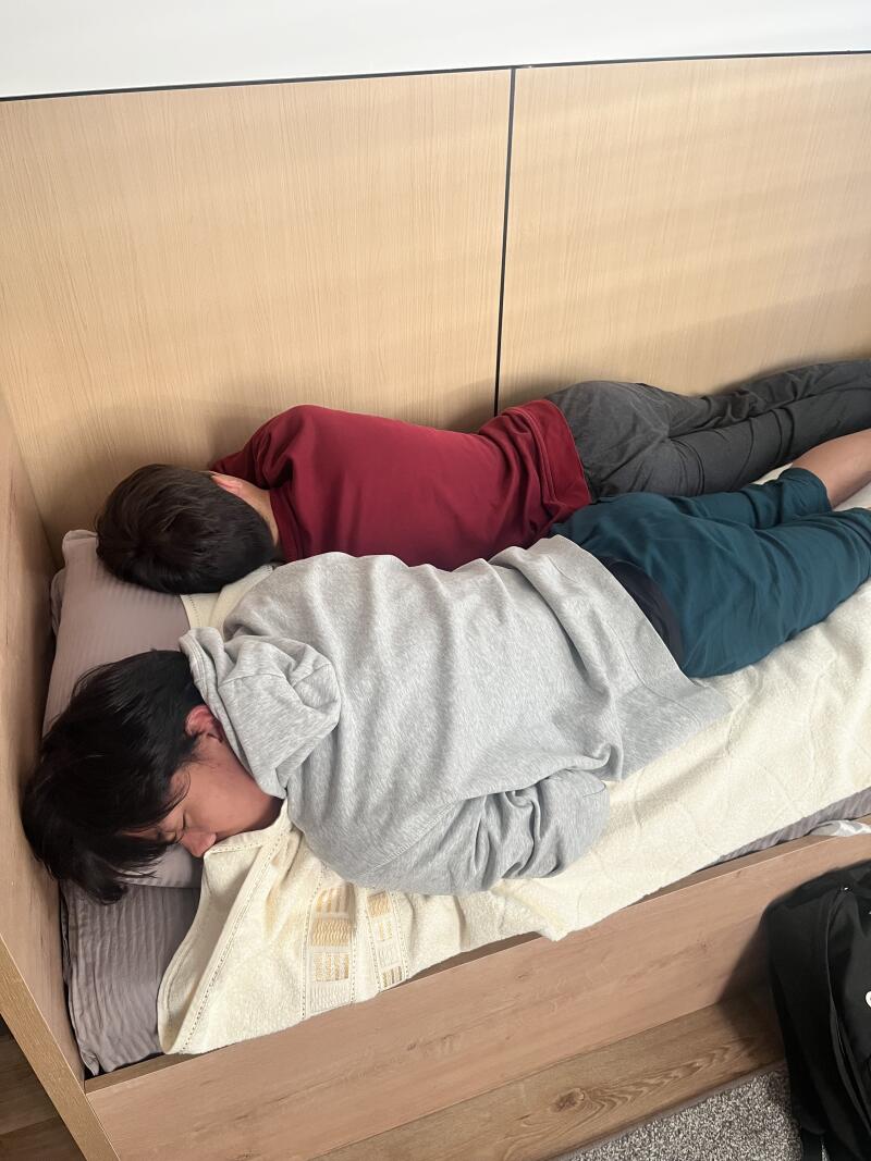Two people are lying on a bed, one wearing a maroon shirt and gray pants, and the other wearing a gray hoodie and blue-green pants.