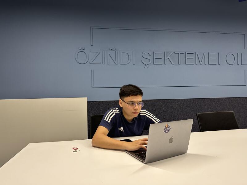A person wearing glasses and an Adidas shirt is sitting at a table using a laptop, with the text "OZINDI ŞEKTEMEI OIL" visible in the background.