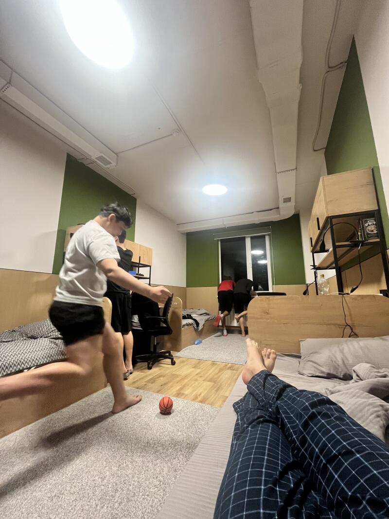 In a dorm room, a person in pajamas lies on a bed while others stand and move around; one person is mid-stride with a small basketball near their foot.