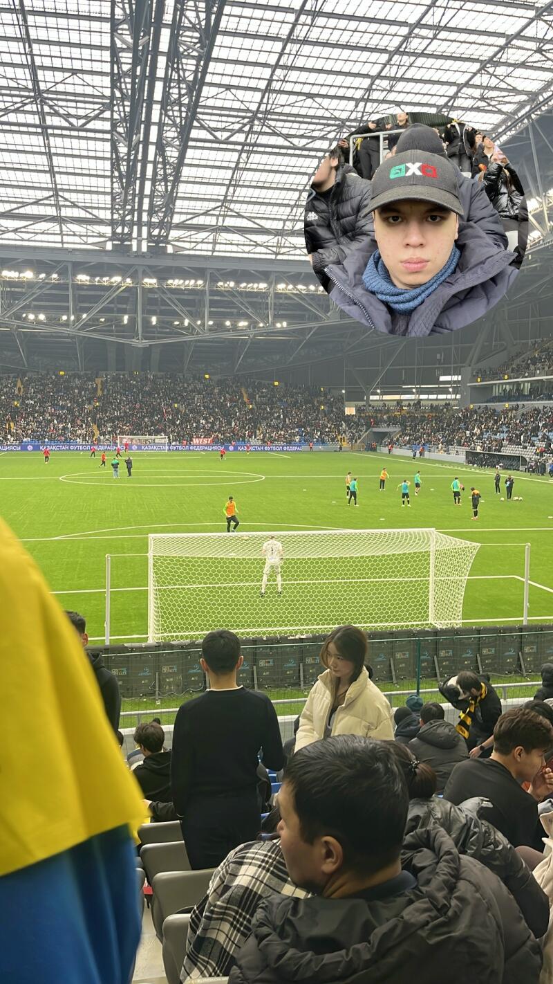 The photo shows a soccer game in a stadium with a person's selfie in the upper right corner. The field is green, and there are players in different color jerseys. There are spectators in the stands.