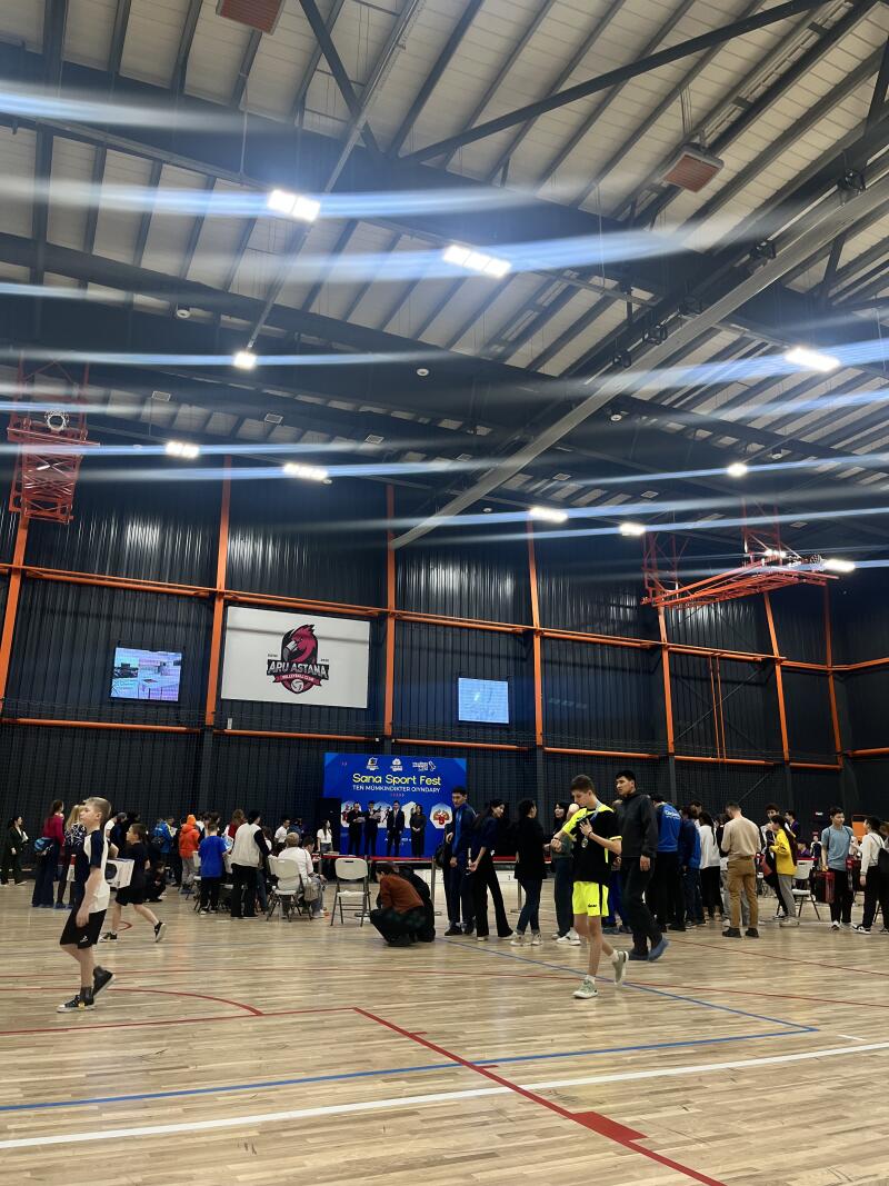 Inside a gymnasium, a crowd gathers near a stage with a banner that reads "Sana Sport Fest". The event is taking place in front of a wall with the logo of "ARU ASTANA" displayed.