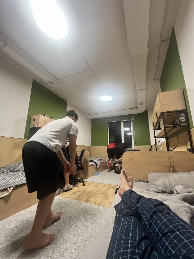 A person is lying in bed wearing blue plaid pajamas, while another person in a white shirt and black shorts stands nearby; in the background, two people are looking out the window.