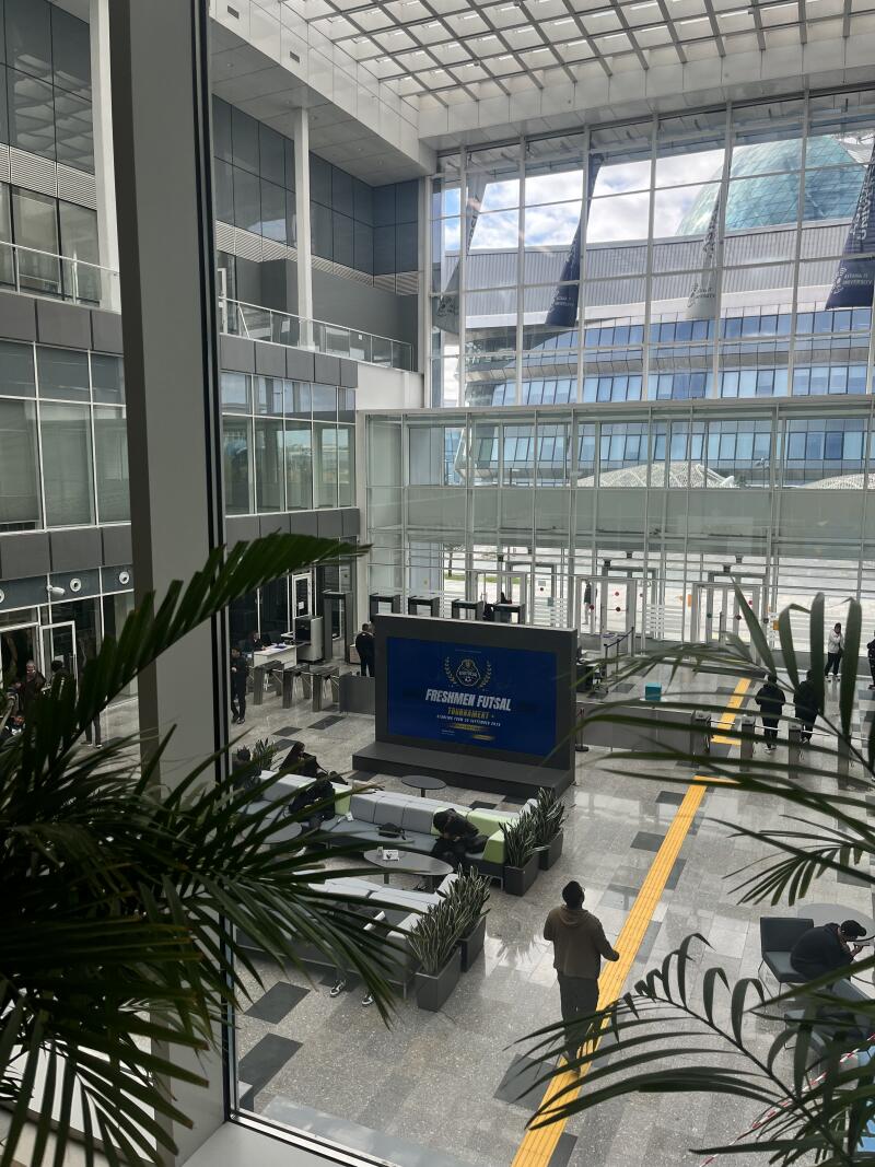 The interior of a modern building features a large screen displaying "Freshmen Futsal Tournament" along with seating areas, plants, and a high glass ceiling. People are seen walking around the space.