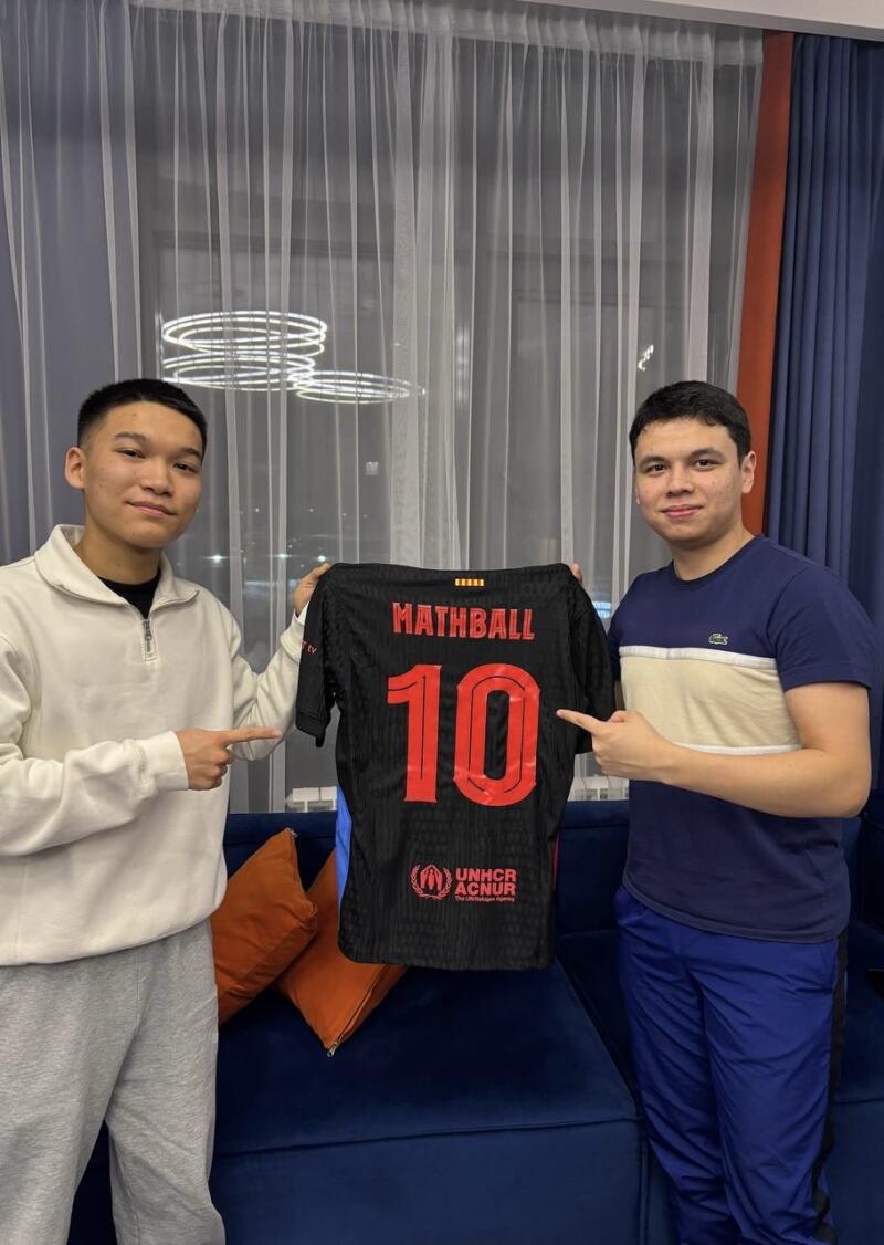Two young men are holding up a black soccer jersey with the name "MATHBALL" and the number 10 in red on the back, along with the UNHCR logo.
