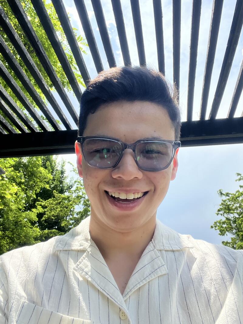 A smiling person wearing glasses and a striped shirt stands under a wooden pergola with greenery visible in the background.