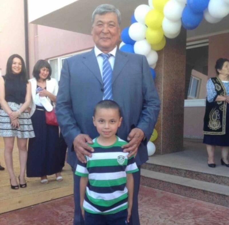 A man in a suit stands with a boy in a striped shirt, with other people and balloons in the background.