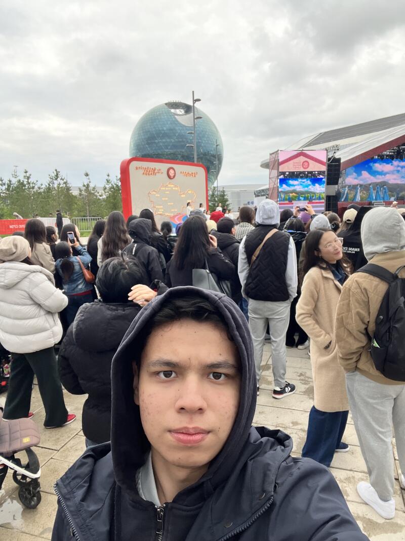 A selfie of a person in a hooded jacket in a crowd of people, with the Nur Alem sphere and a stage visible in the background.