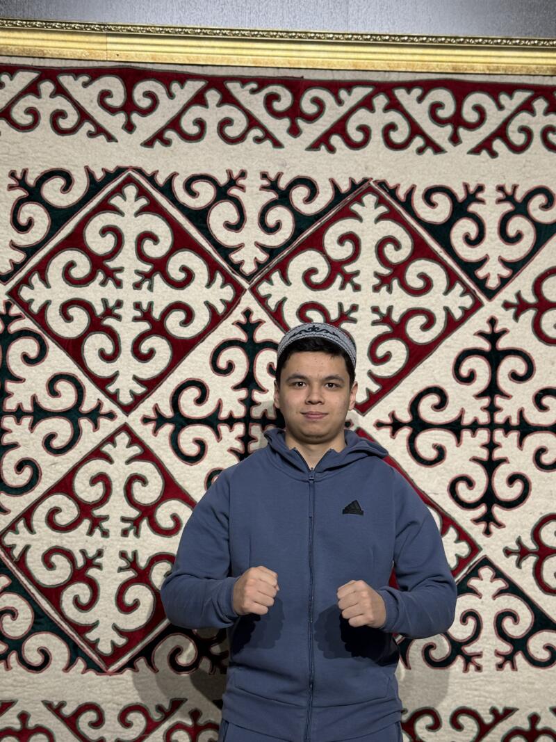 A man wearing a taqiyah and a blue tracksuit stands in front of a wall decoration featuring red and black patterns on a white background.