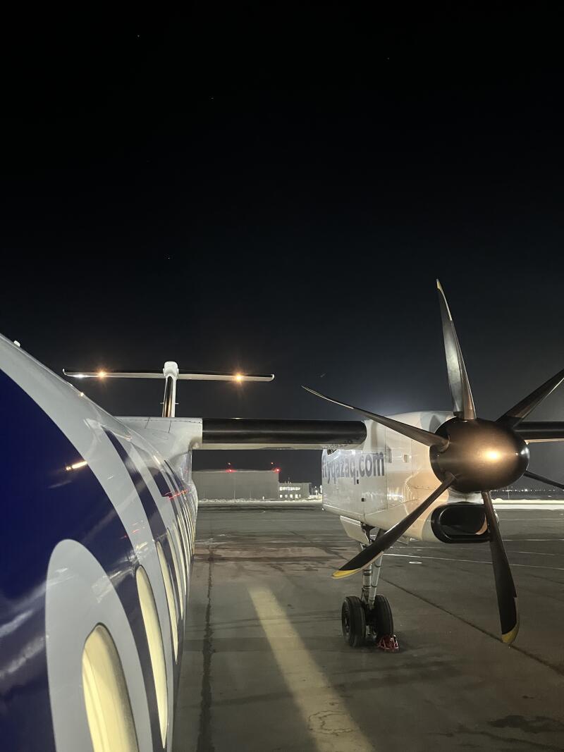 The image shows two FlyArystan airplanes on the ground at night; the closest aircraft has a blue and white design, while the other plane has a visible propeller and the text 'flyqazaq.com' on its side.