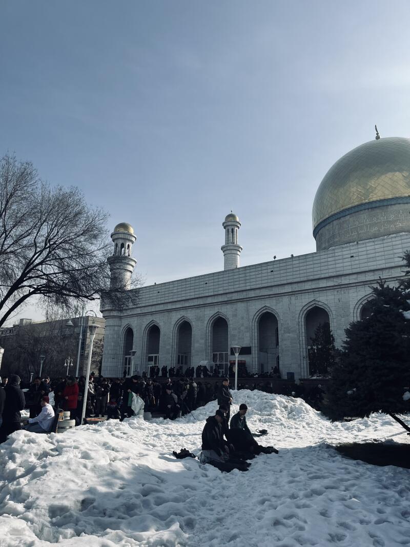 A large group of people are gathered in front of a mosque with a golden dome, some kneeling on the snow-covered ground.
