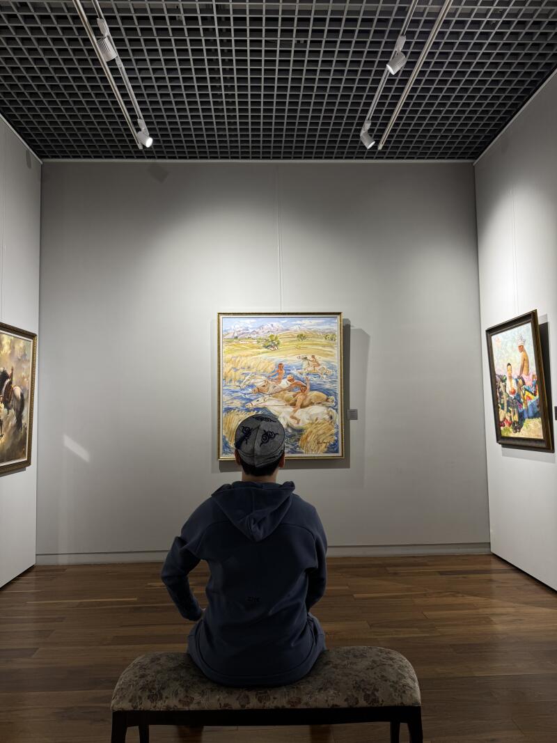 A person wearing a tubeteika and a blue hoodie sits on a bench looking at a painting in an art gallery.