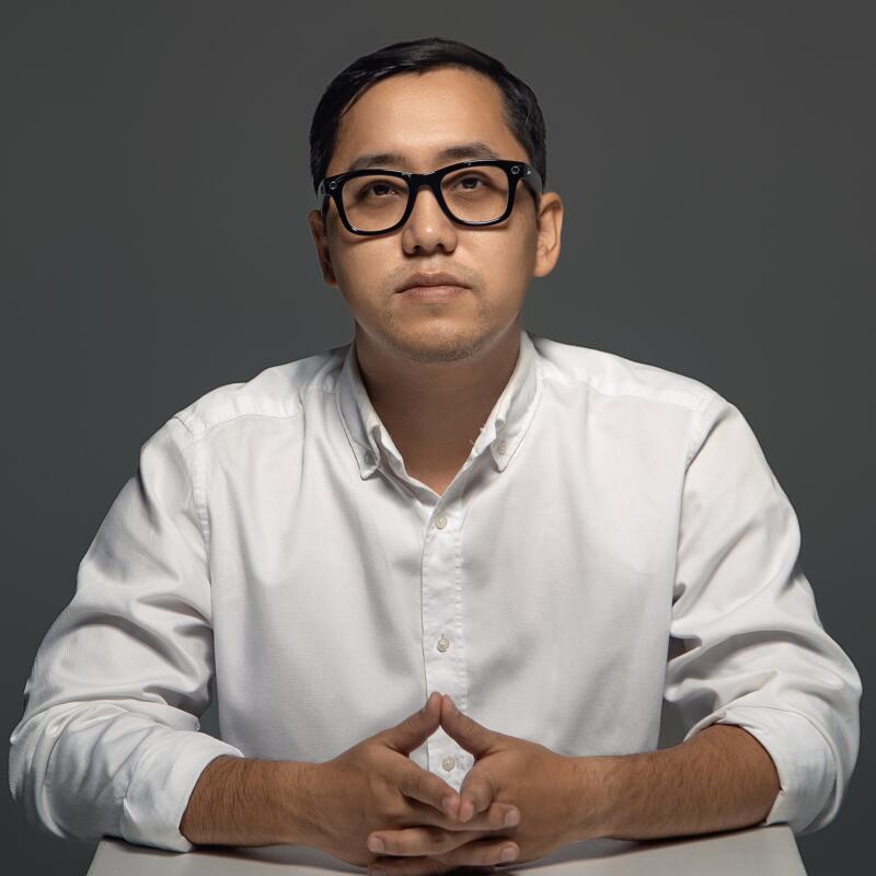 A man wearing black glasses and a white shirt sits at a table with his hands clasped together.