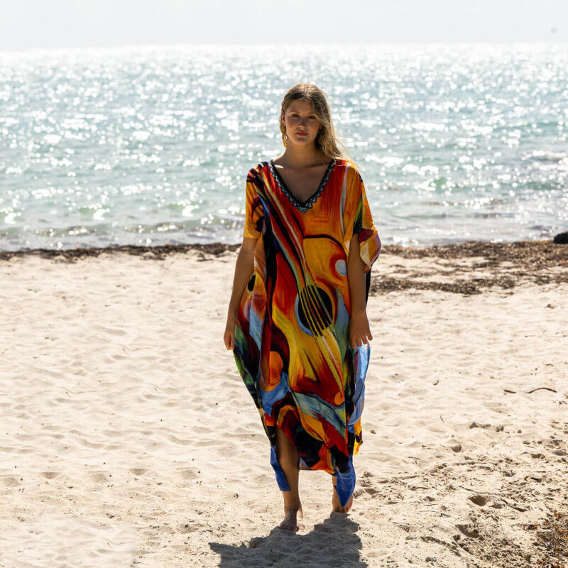 A woman stands on a sandy beach wearing a colorful, guitar-patterned dress, with the ocean visible in the background.