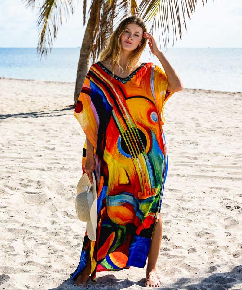 A woman stands on a sandy beach near the ocean, wearing a colorful dress with a guitar print and holding a white hat.