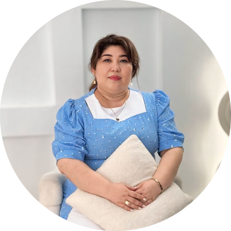 A woman with dark hair is sitting and holding a cream-colored pillow, wearing a blue dress with white trim, and gold jewelry, against a white background.