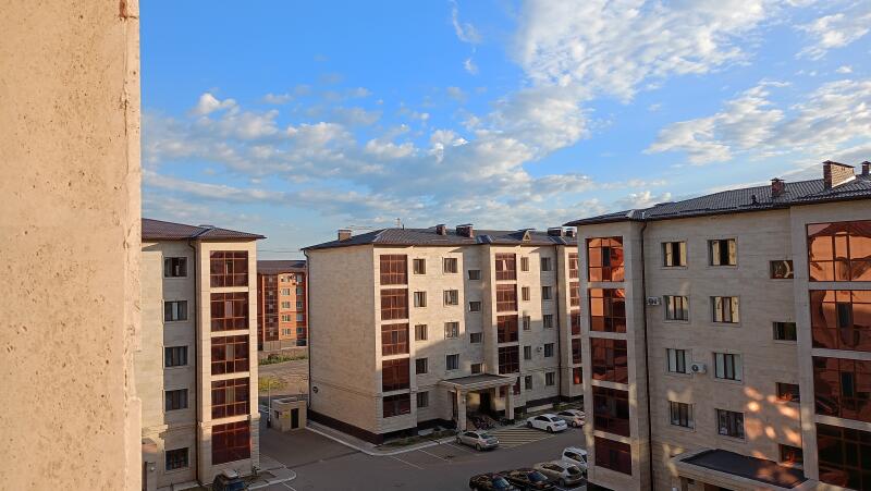 The image shows several multi-story residential buildings with brown roofs and window frames under a blue sky with scattered clouds. Cars are parked in the courtyard.