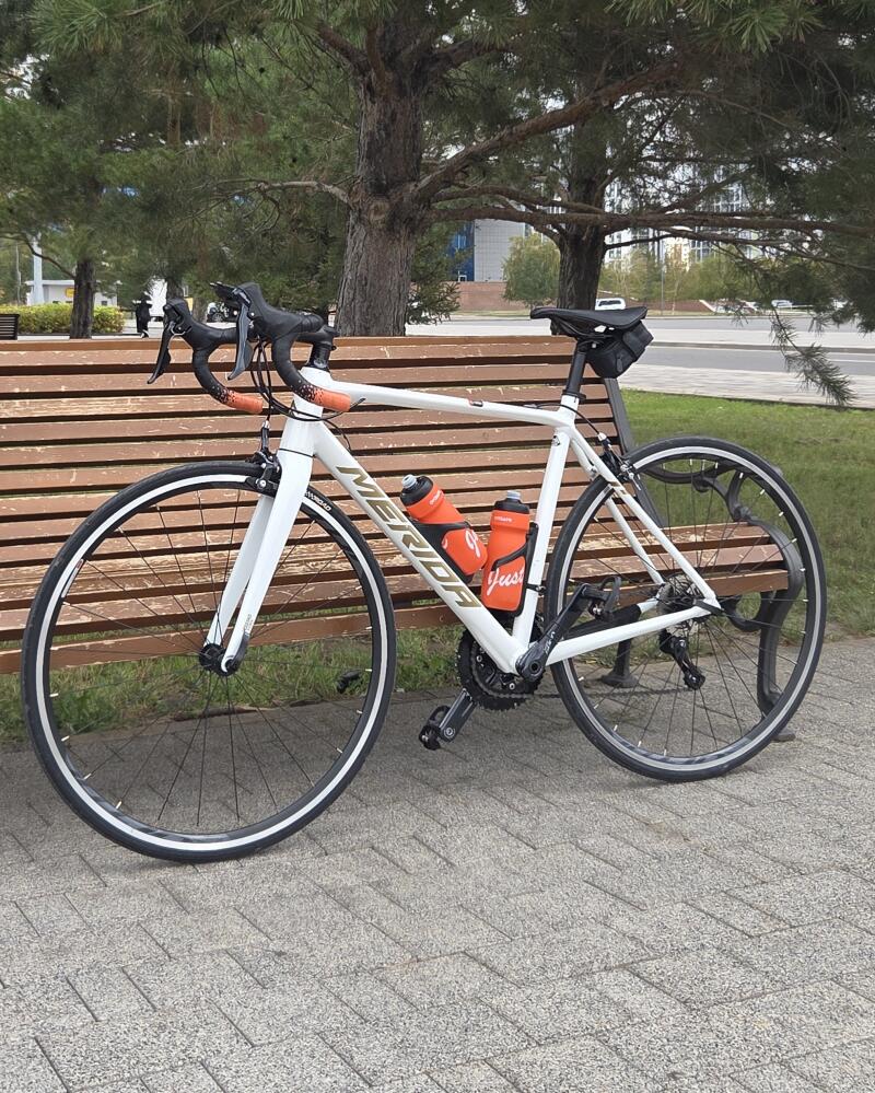 A white Merida road bike with orange accents, two orange water bottles (one with "Just" visible), and a black saddle bag is leaning against a wooden bench on a paved surface, with trees and buildings in the background.