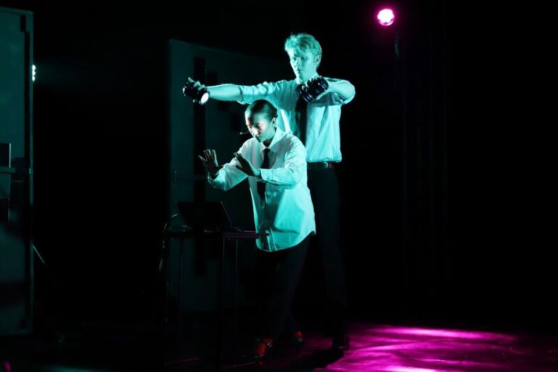 Два человека в белых рубашках и черных галстуках выступают на сцене с темным освещением; один держит в руках фонари, другой стоит возле ноутбука.