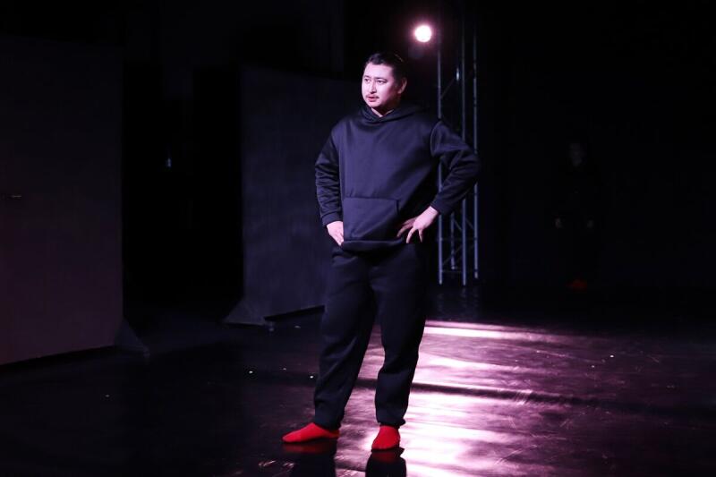 Мужчина стоит на сцене в черном худи и штанах, в красных носках или тапочках, освещенный прожектором.