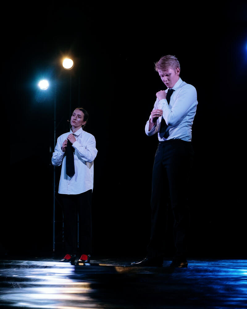 Два человека стоят на сцене, поправляя галстуки и рукава, одетые в белые рубашки на пуговицах и черные брюки.