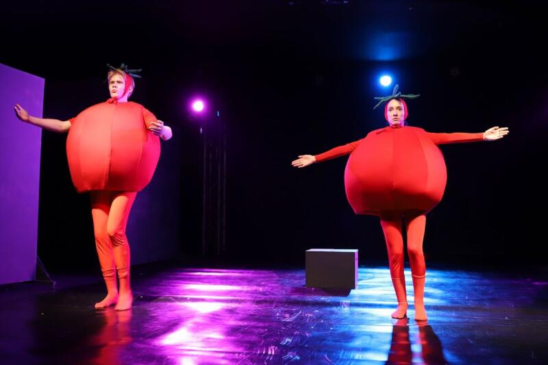 Два исполнителя в красных костюмах помидоров стоят на сцене, вытянув руки.