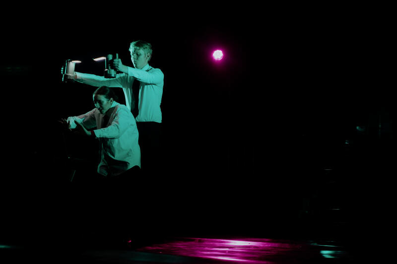 Два исполнителя, мужчина и женщина, освещенные зелеными и розовыми сценическими огнями, запечатлены на темном фоне; мужчина, одетый в белую рубашку и галстук, держит и направляет два осветительных прибора, в то время как женщина, также в белой рубашке, жестикулирует руками в, казалось бы, оборонительной позе.