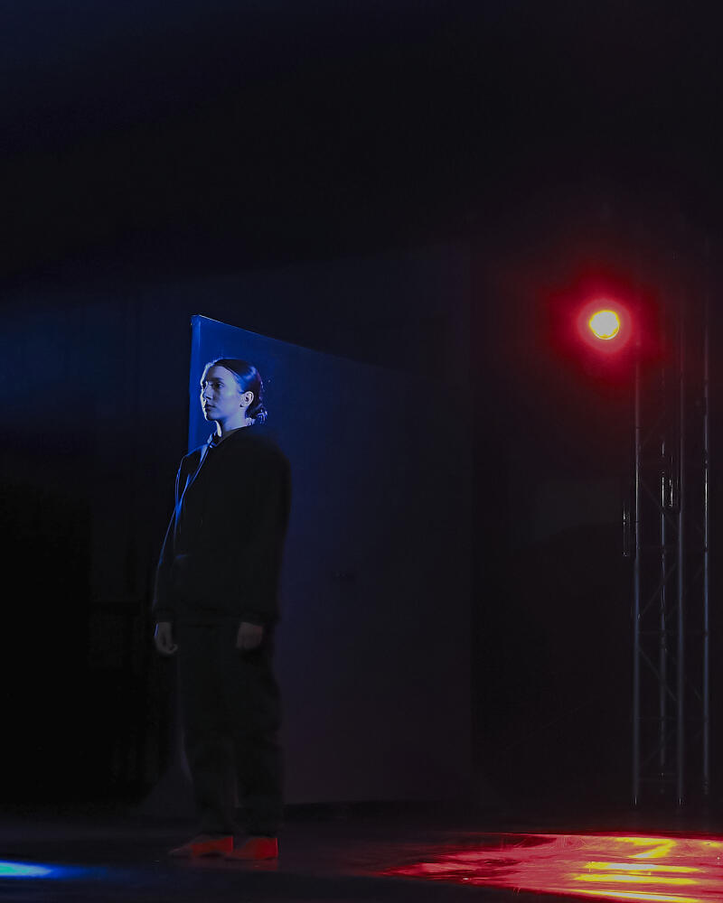 Женщина в темном наряде стоит на сцене, освещенной синим и красным светом, справа видна металлическая конструкция.